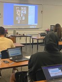 Photo of students working on Chromebooks in the classroom and Zoom students projected on an electronic whiteboard.